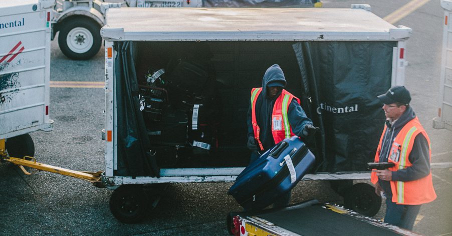 two baggage handlers move bags from a cart to a conveyor belt